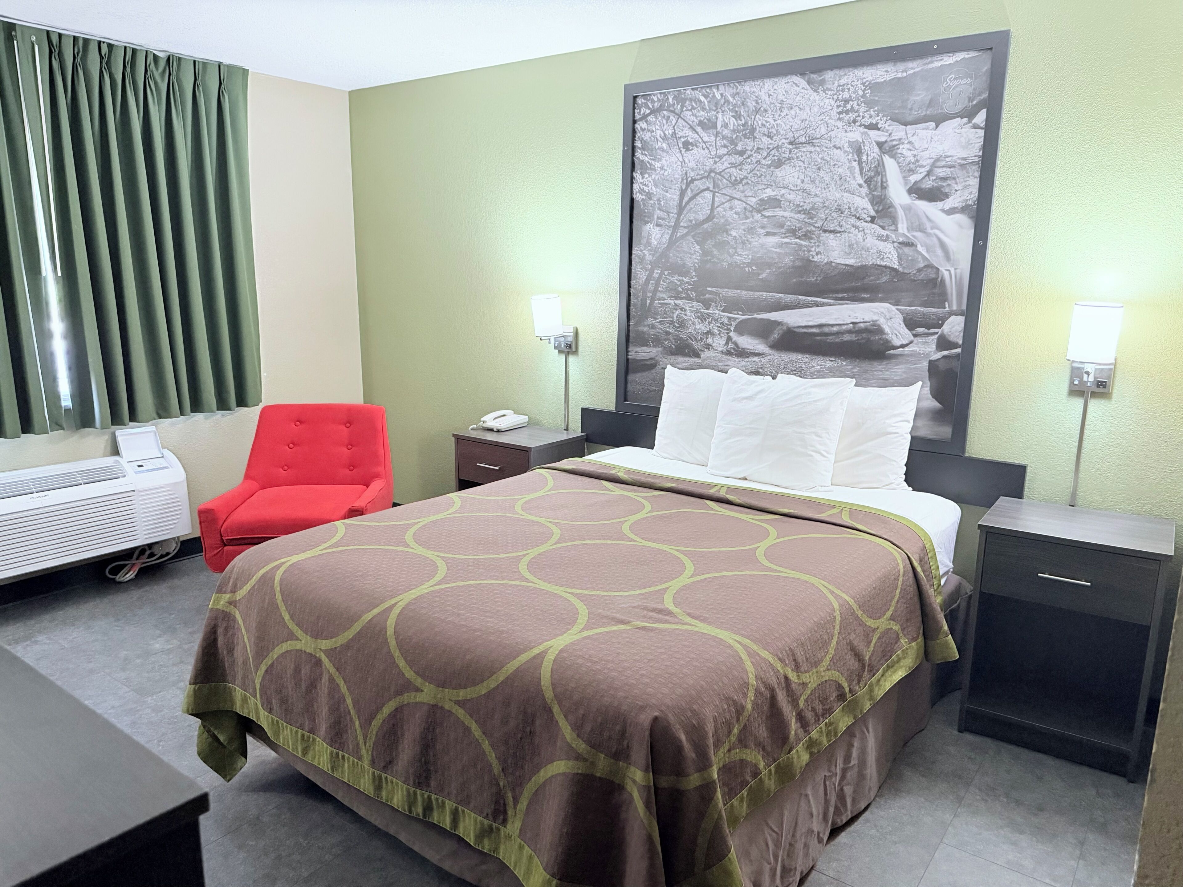 A hotel room with a bed, a red armchair, a desk, a nightstand, a wall-mounted air conditioner, and a large framed picture on the wall.