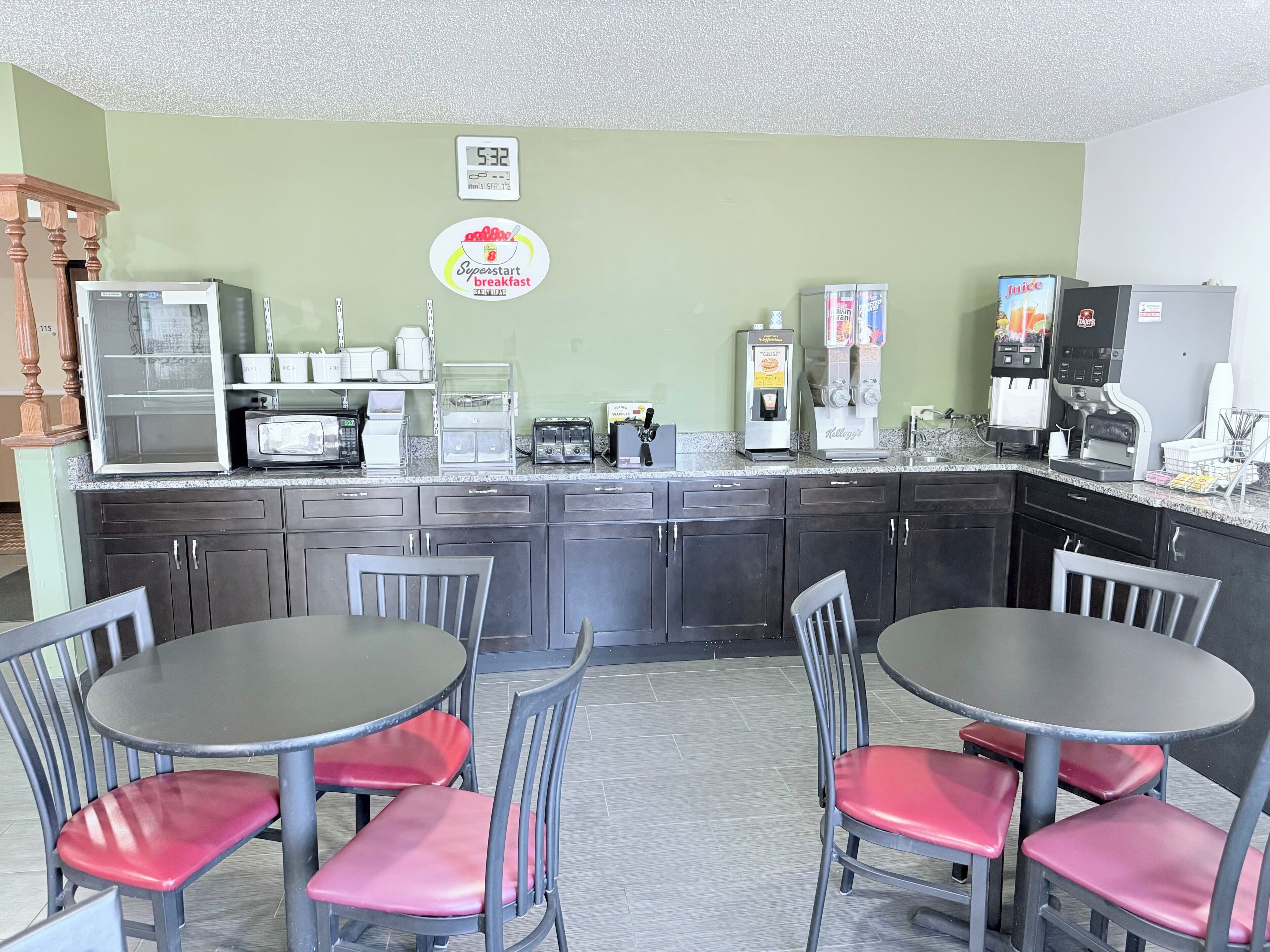 A kitchen with a clock, a Superstart breakfast sign, a microwave, toaster, coffee machine, and a juice dispenser.