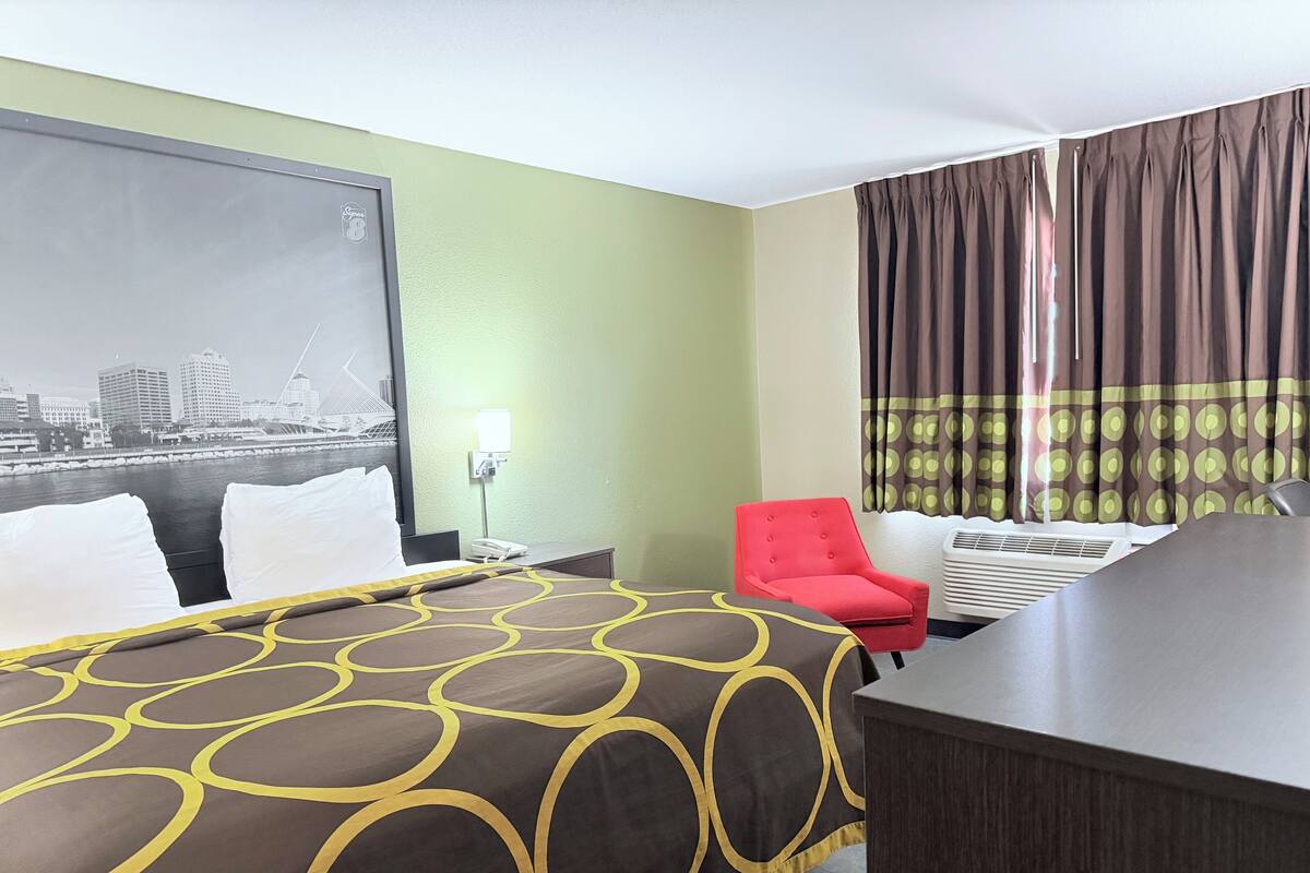 Hotelkamer met een bed, bureau, stoel, nachtkastje, lamp en raam met gordijnen.