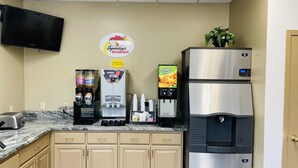 Een kitchenette met een koffieapparaat, een waterdispenser, een koelkast en een aan de muur bevestigde televisie.