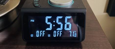 Een digitale klok toont 17:56, met een temperatuur van -12 graden Celsius, en een aan/uit-knop op een houten ondergrond.