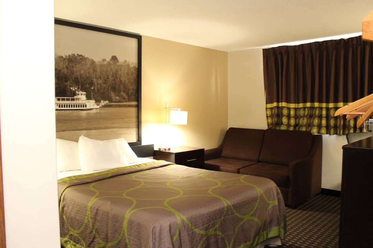 Une chambre d’hôtel avec un lit, un bureau, une chaise, une lampe et une grande fenêtre avec des rideaux.