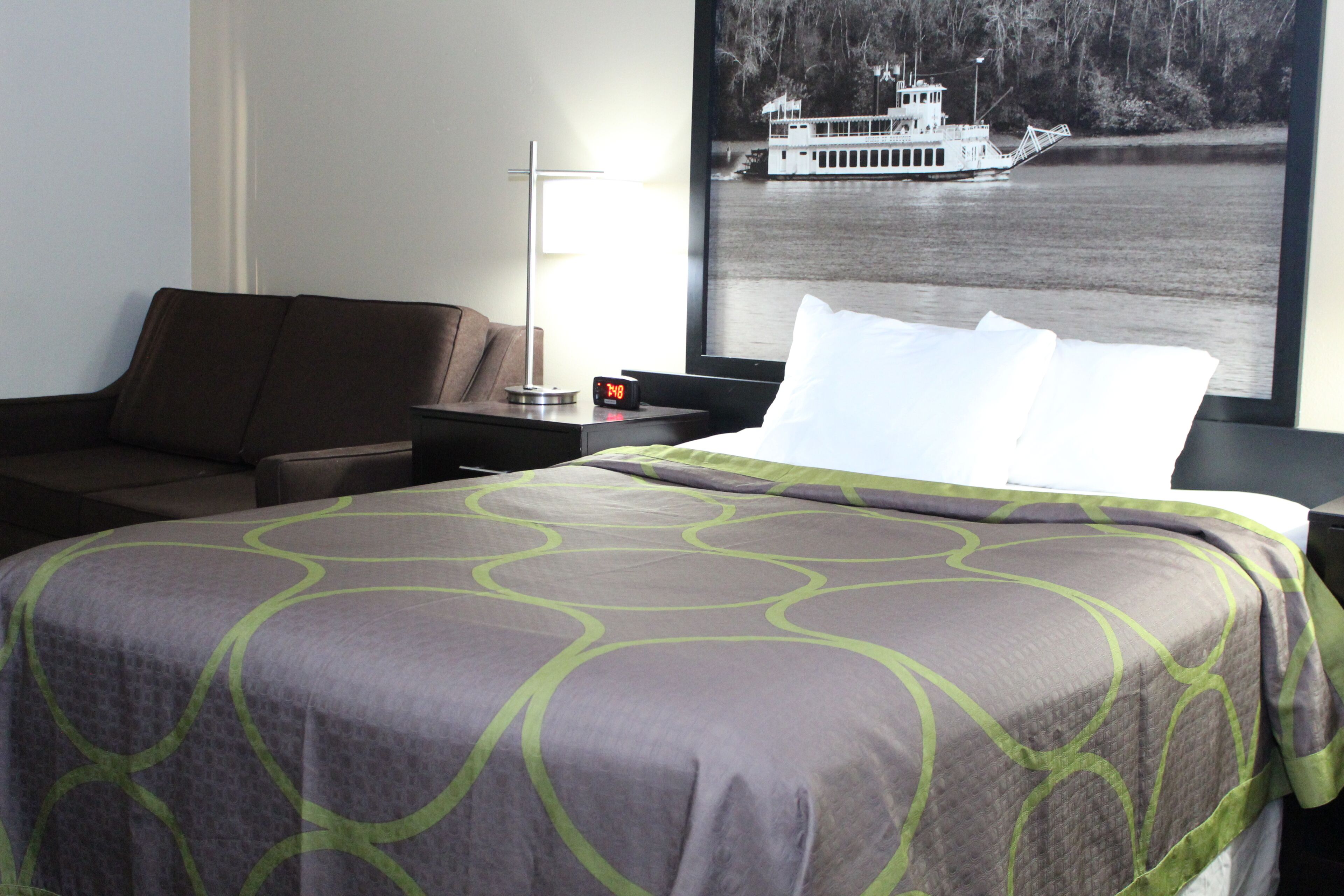 A hotel room with a bed, a nightstand, a lamp, and a poster of a boat on a lake.