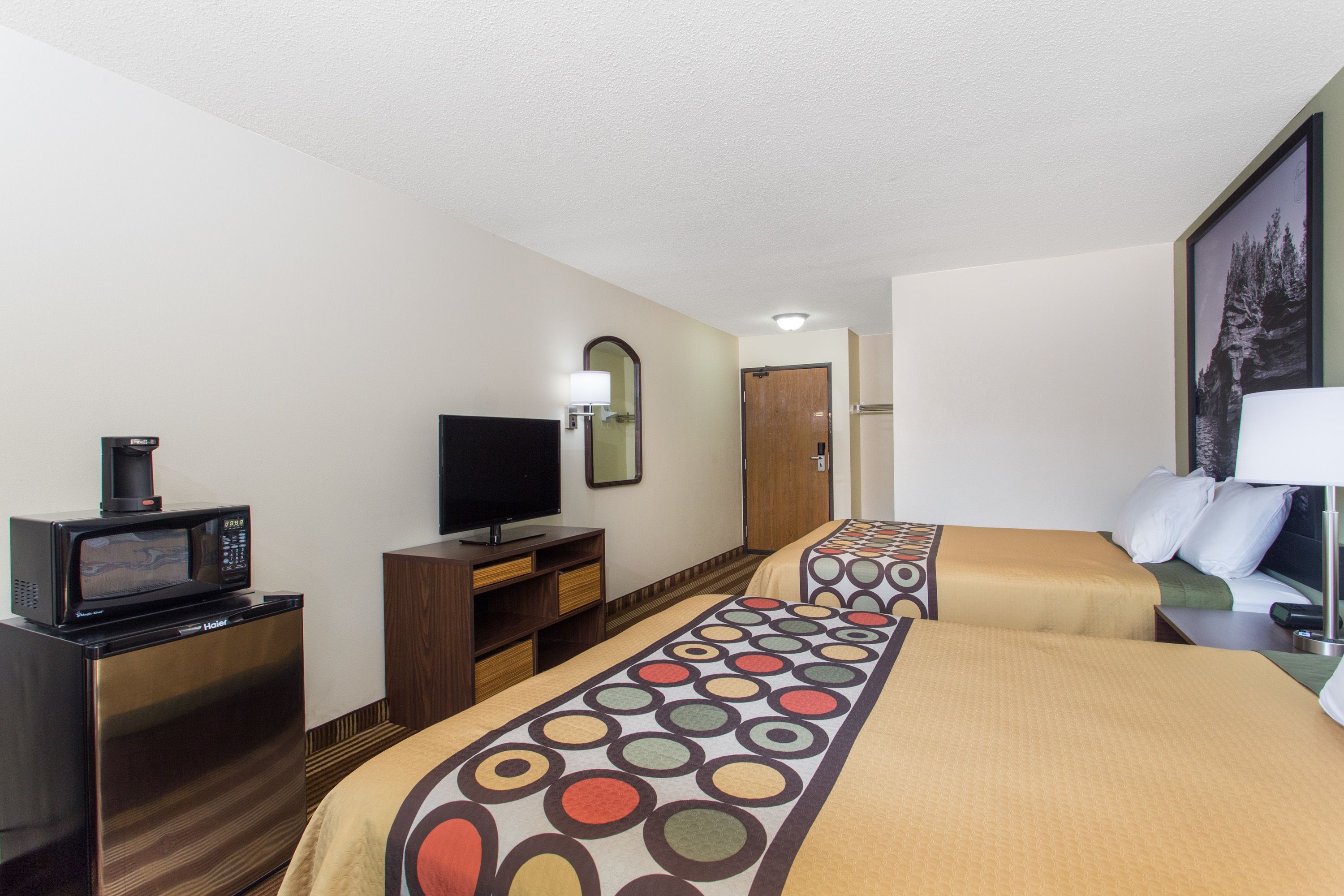 A hotel room with a kitchenette, a television, a bed with a patterned bedspread, and a painting on the wall.