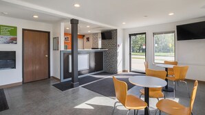 A modern indoor space with a reception desk, a flat-screen TV, and a seating area with round tables and chairs. - Motel 6 Marshalltown, IA (Marshalltown)