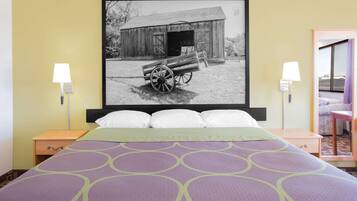 A hotel room with a bed, two bedside tables, a nightstand, a framed picture of a barn and wagon, and a window with curtains.
