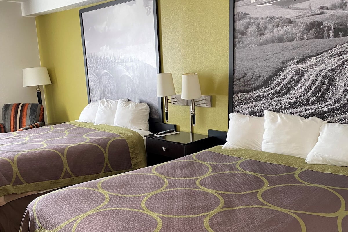 A hotel room with two beds, a large window, and a wall-mounted lamp.
