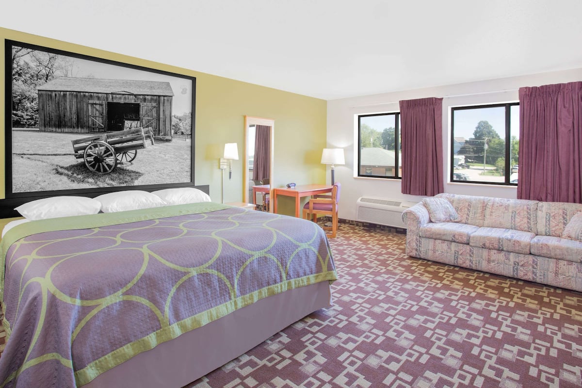 A hotel room with a large bed, a sofa, a desk, a chair, a window with curtains, and a wall-mounted picture.