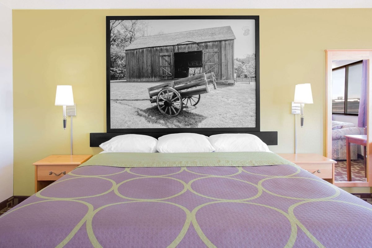 A hotel room with a bed, two bedside lamps, a large framed picture of a barn and a cart, and a wooden nightstand.