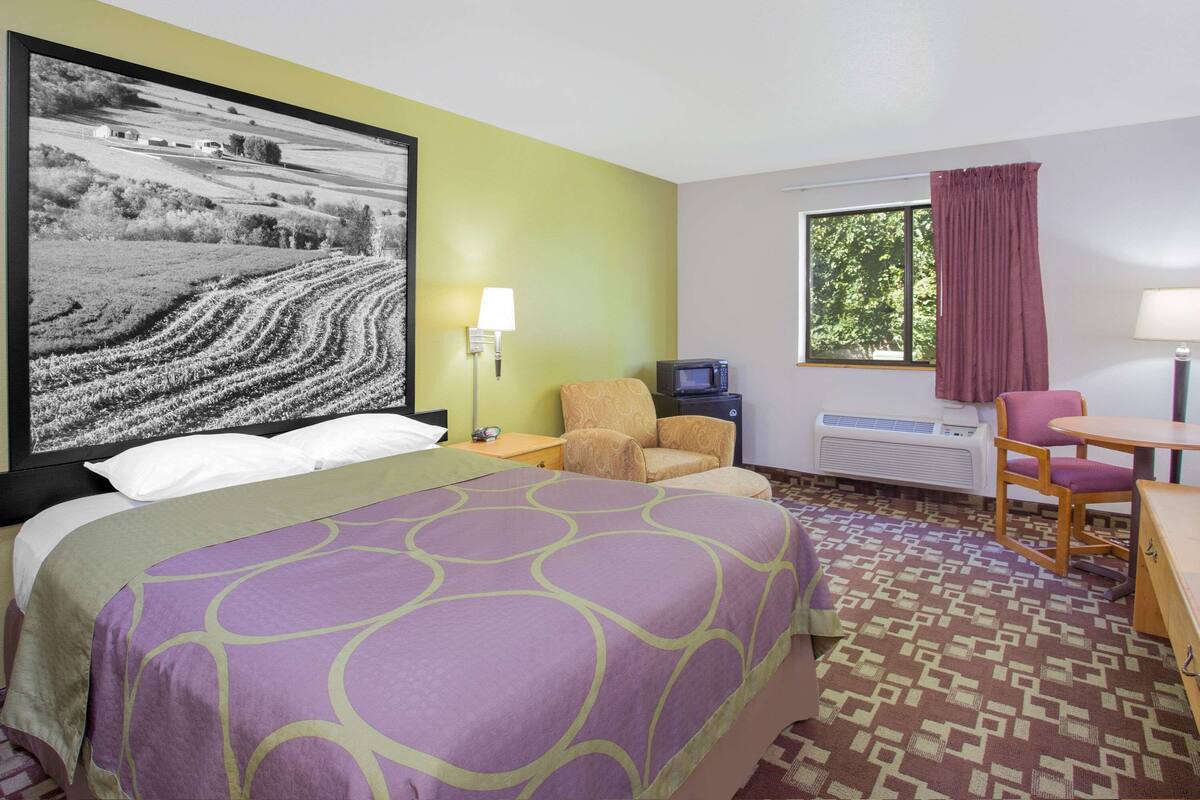 A hotel room with a bed, a chair, a table, a TV, and a window with curtains.