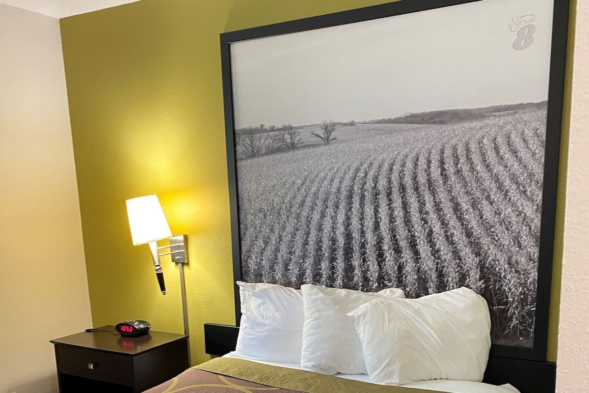 A hotel room with a bed, a nightstand with a lamp, and a large framed picture of a field.