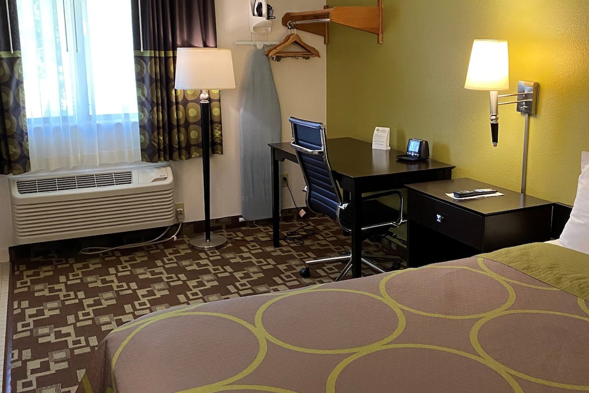 A hotel room with a bed, desk, chair, ironing board, and a wall-mounted lamp.