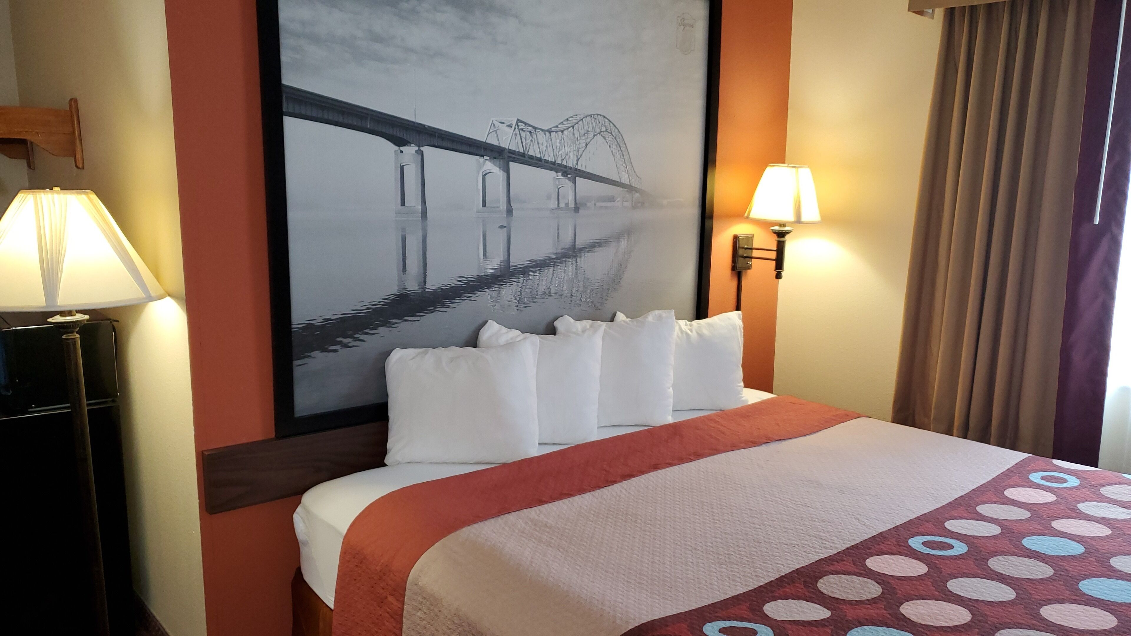 Ein Hotelzimmer mit einem großen Bett, zwei Nachttischlampen, einem am Wand montierten Fernseher und einem Fenster mit Vorhängen.
