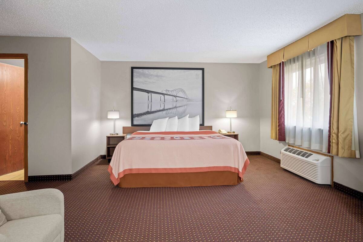 A hotel room with a large bed, two bedside tables with lamps, a large window with curtains, and a wall-mounted air conditioner.