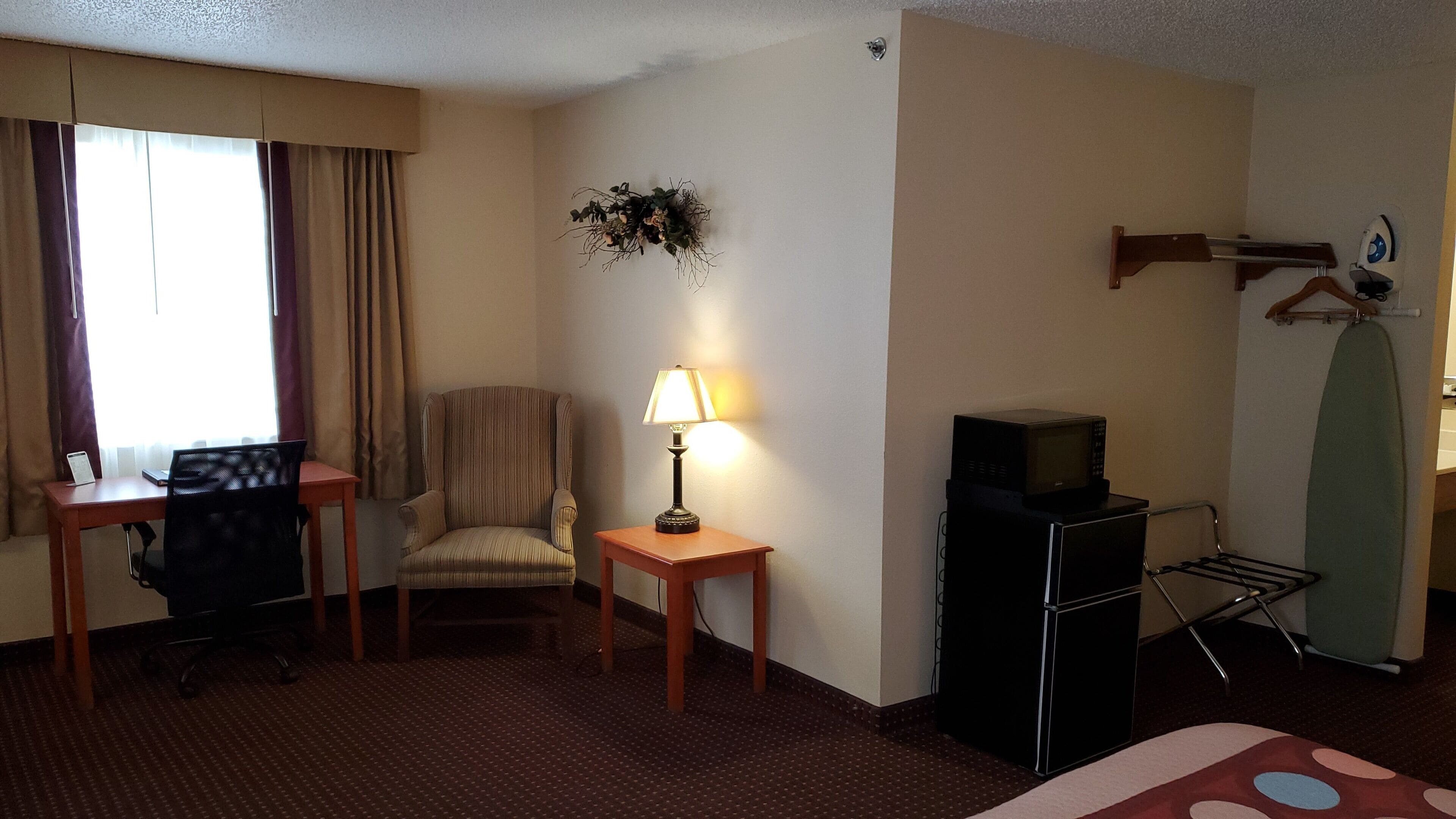 A hotel room with a desk, chair, lamp, TV, microwave, ironing board, and a bed with a polka dot bedspread.