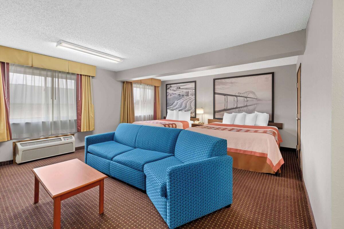 A hotel room with a blue sofa, a wooden table, a bed with a peach-colored bedspread, a nightstand, a lamp, and a large landscape painting on the wall.