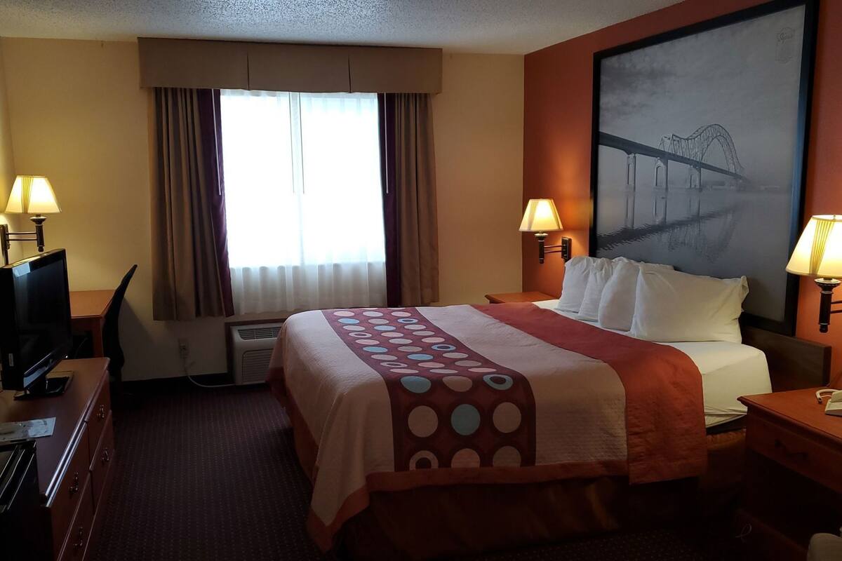 A hotel room with a bed, two bedside lamps, a TV, a desk, and a large window with curtains.