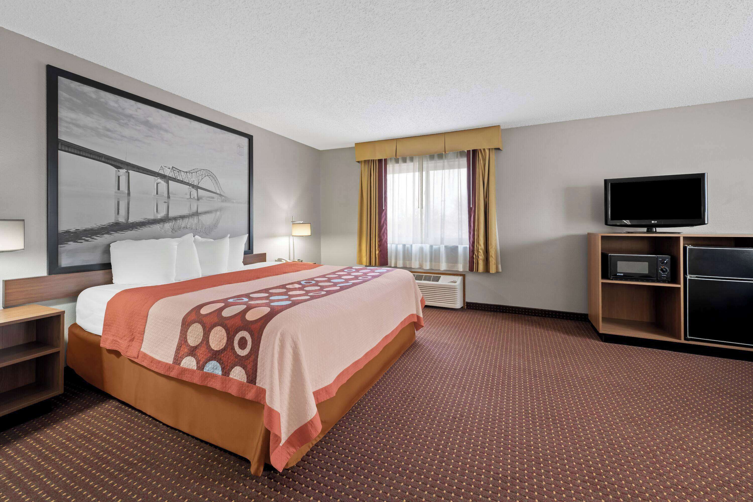Ein Hotelzimmer mit einem großen Bett, einem Flachbildfernseher, einem Schreibtisch mit Lampe und einem großen Fenster mit Vorhängen.