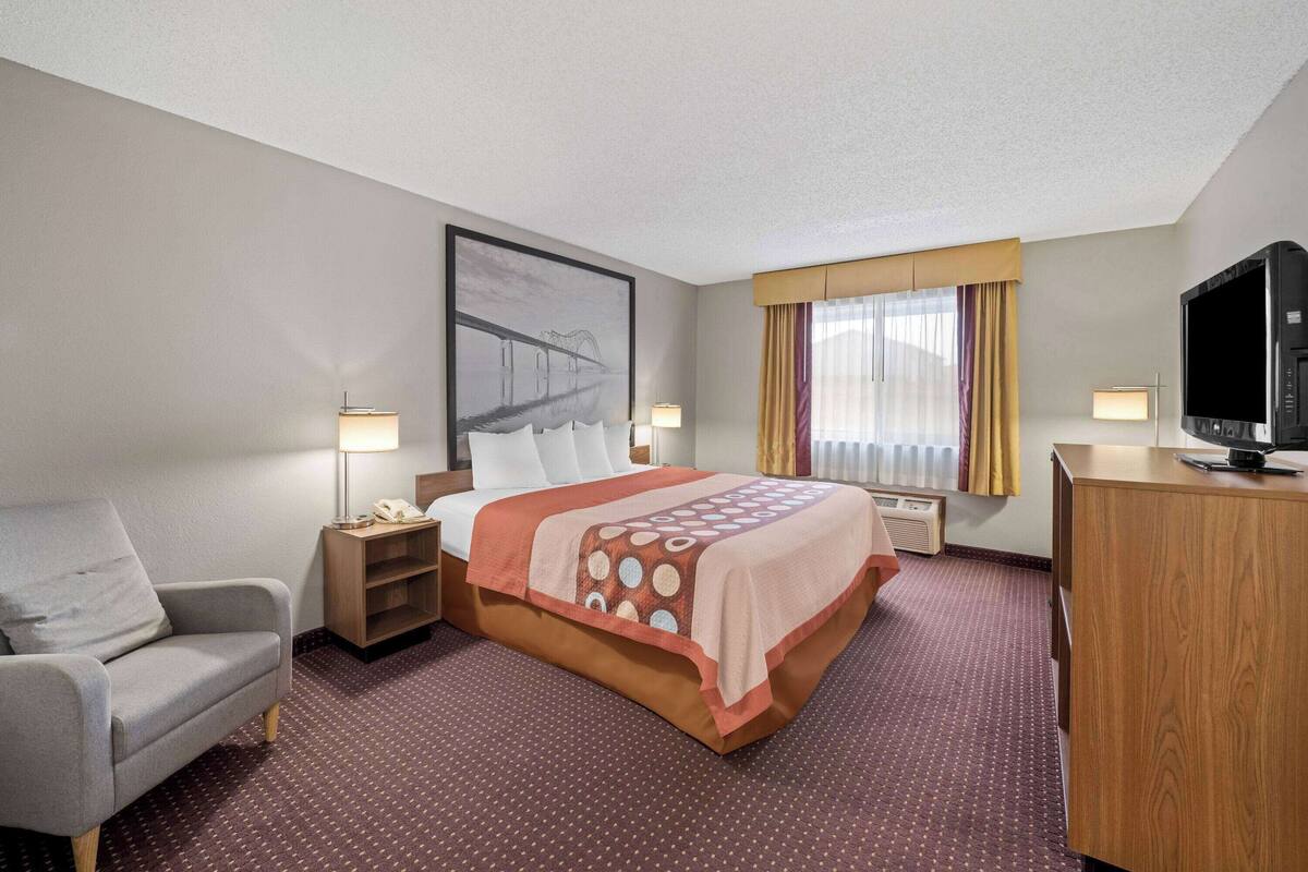 A hotel room with a bed, a desk, a chair, a television, and a painting on the wall.