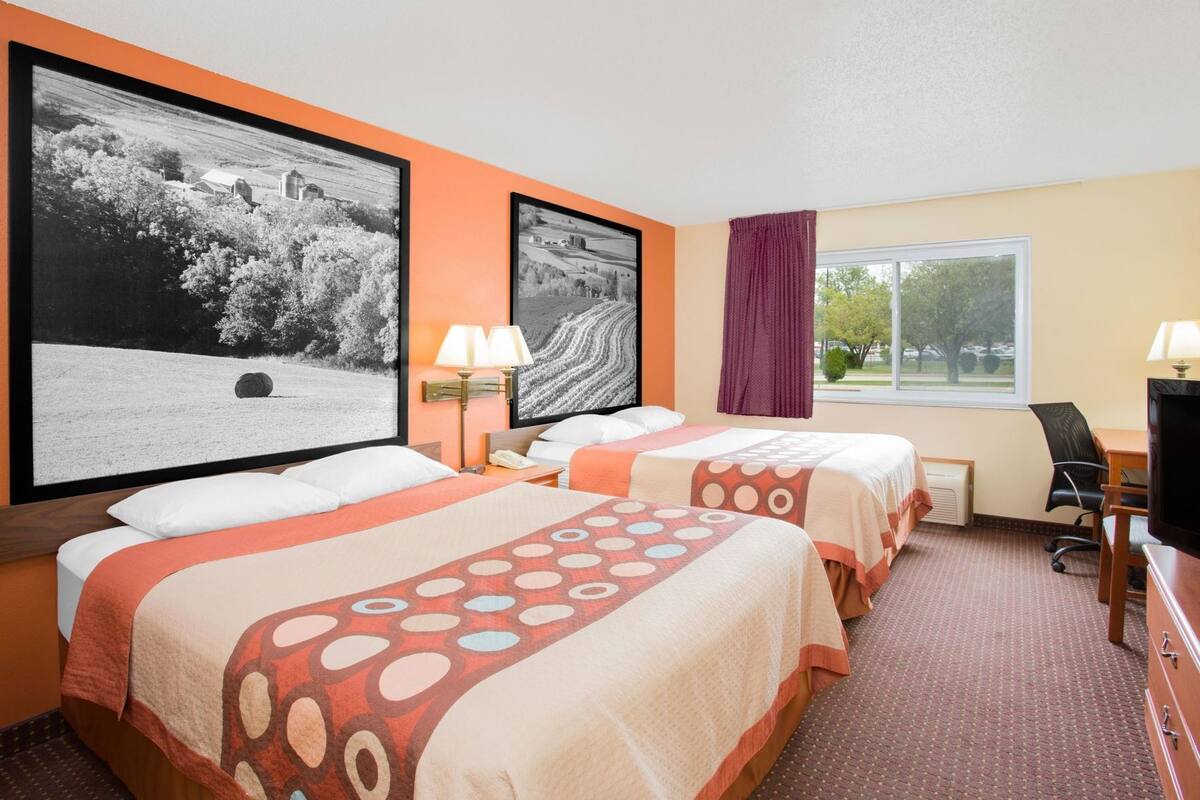 A hotel room with two beds, a desk, a chair, a TV, and two large landscape photographs on the wall.