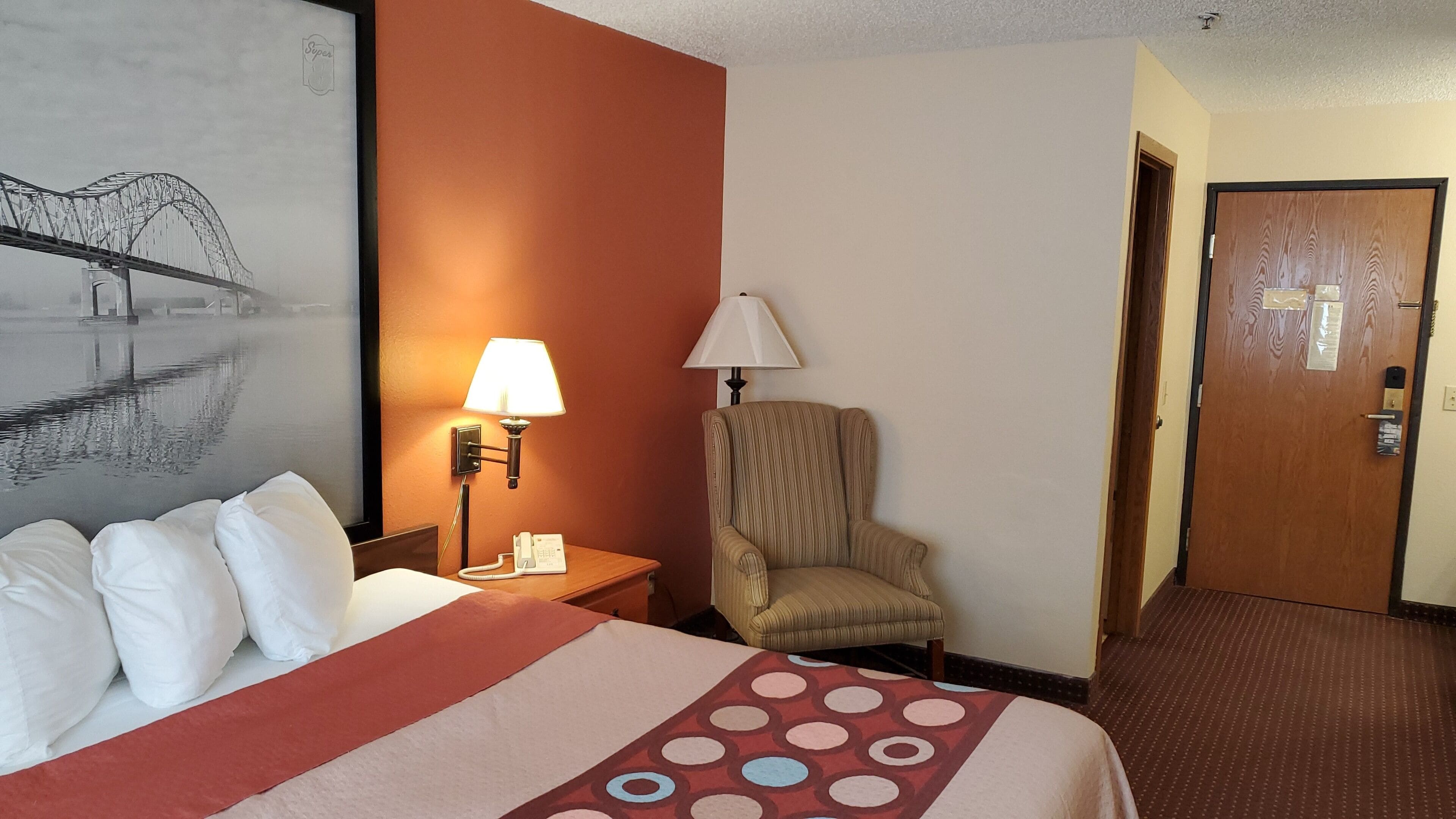 Ein Hotelzimmer mit Bett, Stuhl, Lampe, Telefon und Tür.