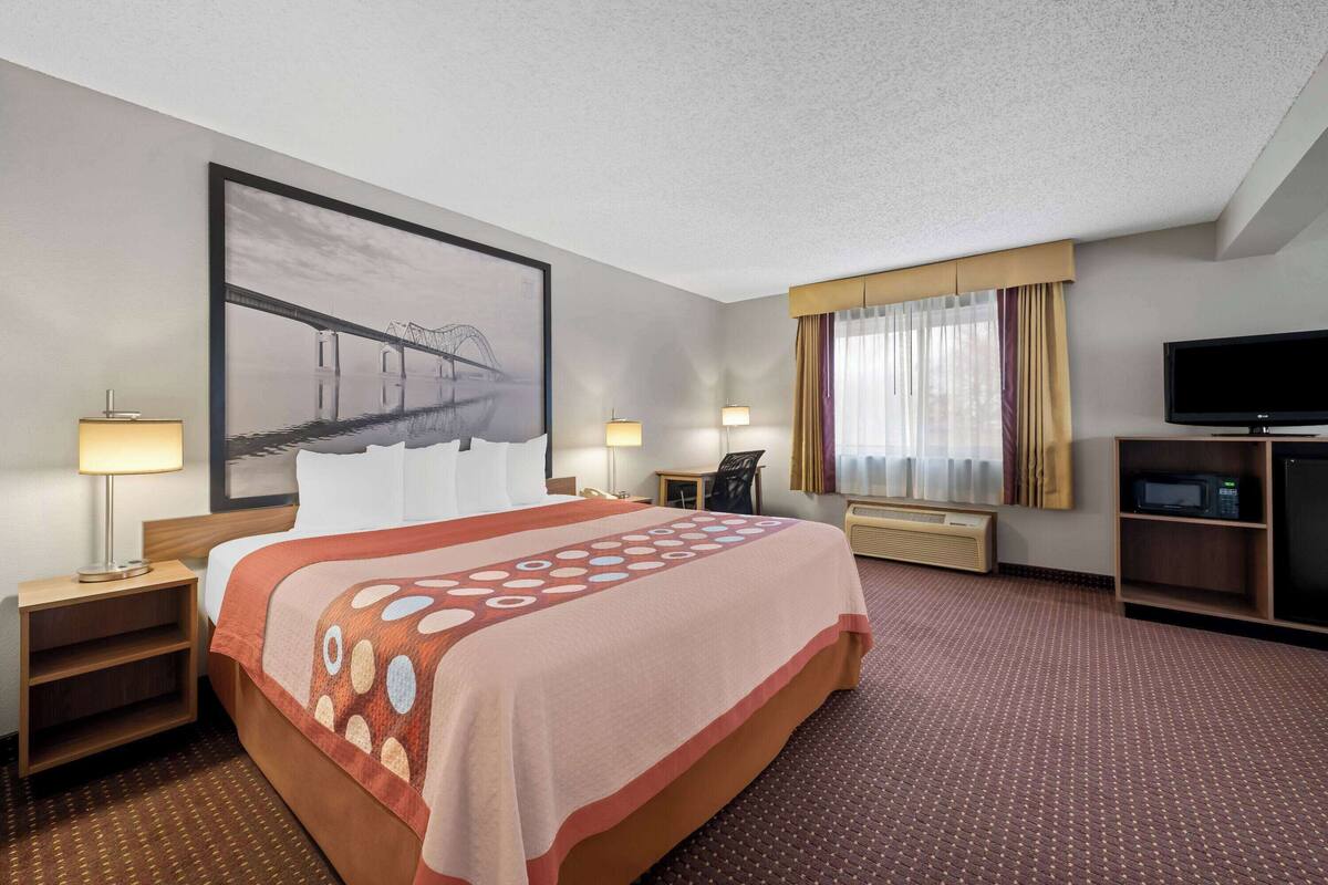 A hotel room with a large bed, a flat-screen TV, a desk with a chair, a lamp, and a painting of a bridge.