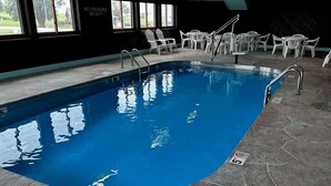 Una piscina interior con agua cristalina, rodeada de sillas y mesas de plástico blanco, y un cartel de socorrista en la pared.
