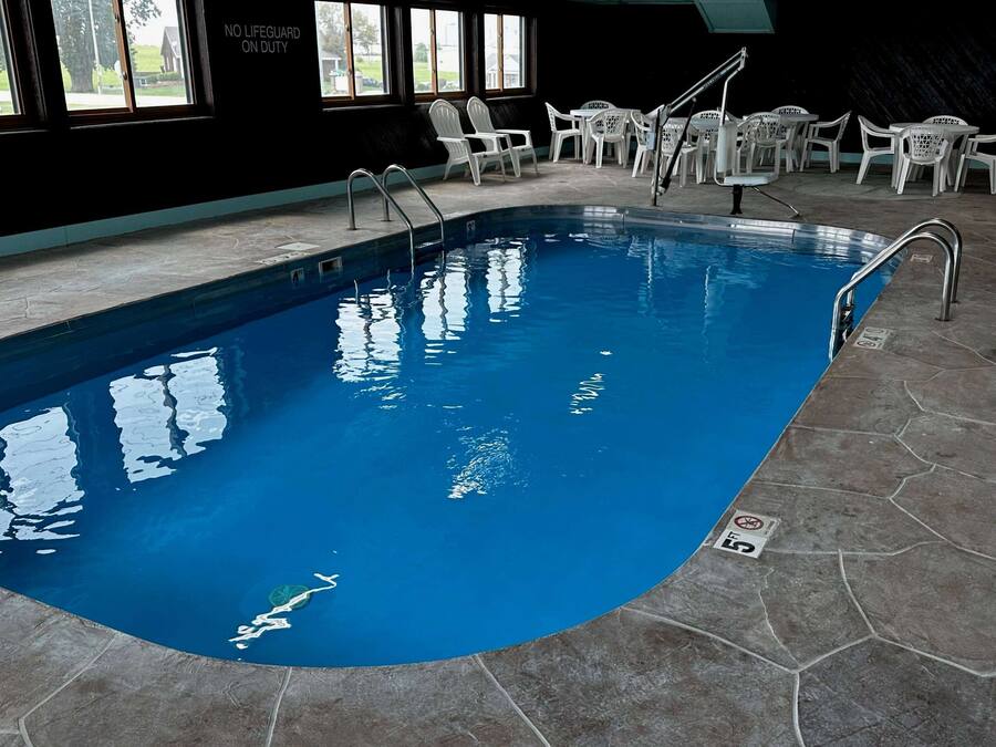 An indoor swimming pool with clear water, surrounded by white plastic chairs and tables, and a lifeguard sign on the wall.