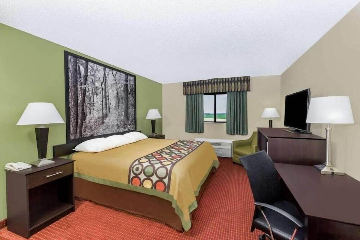 A hotel room with a bed, two bedside tables with lamps, a desk with a chair, a television, and a large window with curtains.