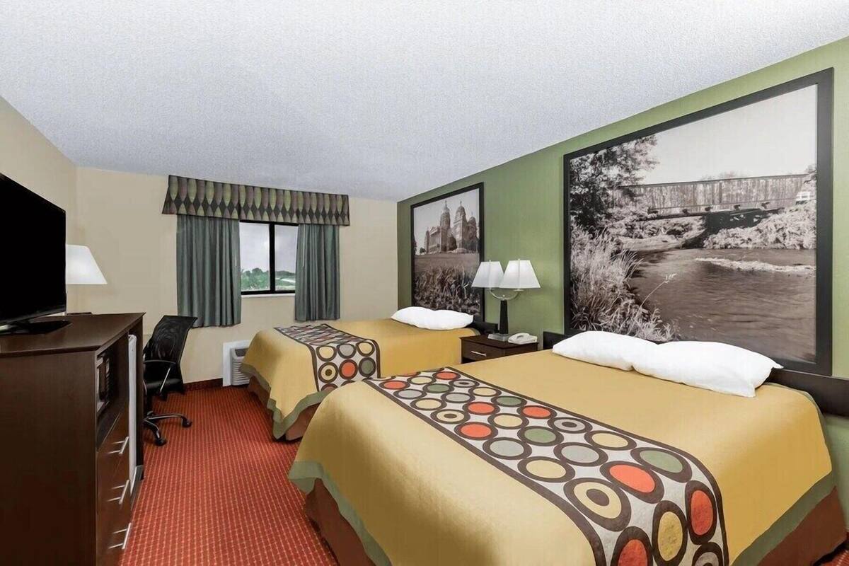 A hotel room with two beds, a desk, a TV, a window with curtains, and a large picture on the wall.