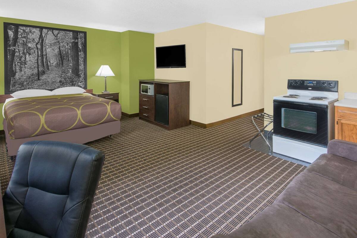 A hotel room with a bed, a desk, a chair, a television, a microwave, a stove, and a large painting on the wall.