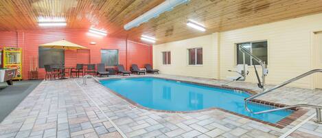 Una piscina interior con suelo de baldosas, techo de madera y una zona de estar junto a la piscina con sillas y una mesa.