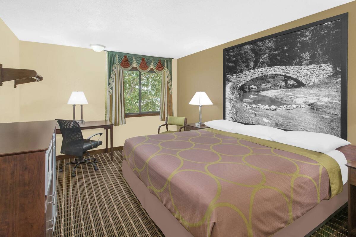 A hotel room with a bed, two chairs, a desk, a lamp, a window with curtains, and a large framed picture on the wall.