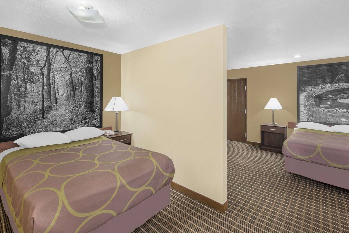 A hotel room with two beds, a large forest-themed painting on the wall, a patterned carpet, and two bedside lamps.