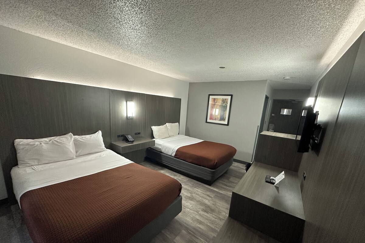 A modern hotel room with two beds, a bedside table with a phone, a small desk with a chair, a wall-mounted TV, and a framed picture on the wall.
