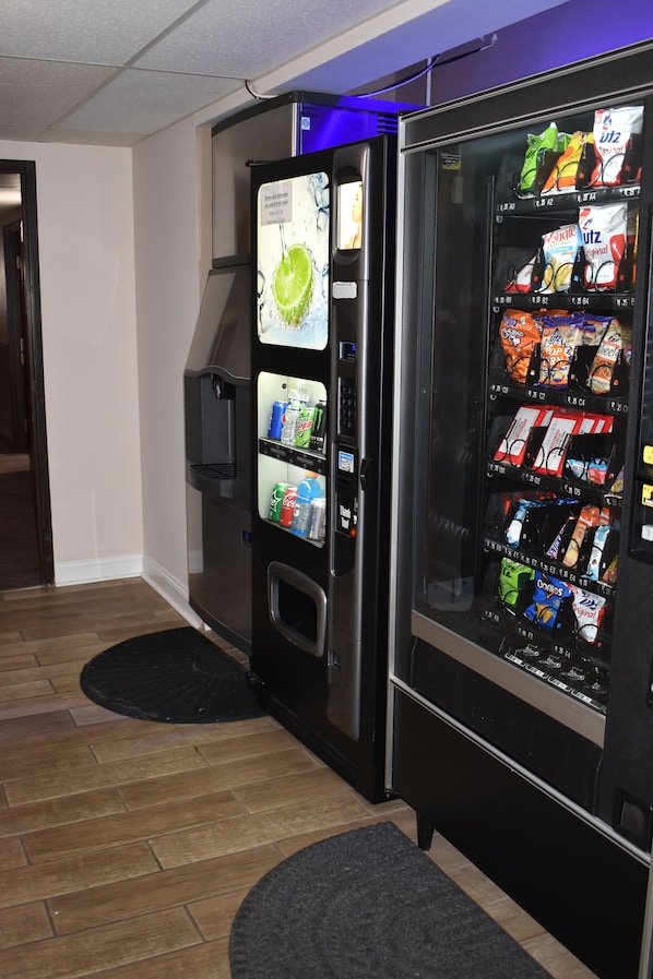 Máquina de vendas com diversos snacks e bebidas, situada em um corredor com piso de madeira e tapete preto.