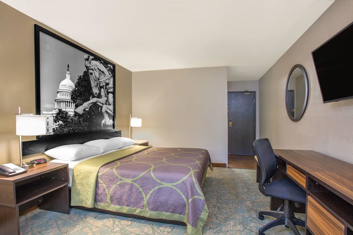 Une chambre d’hôtel avec un lit, un bureau, une chaise, une télévision, un tableau représentant le Capitole et une porte.