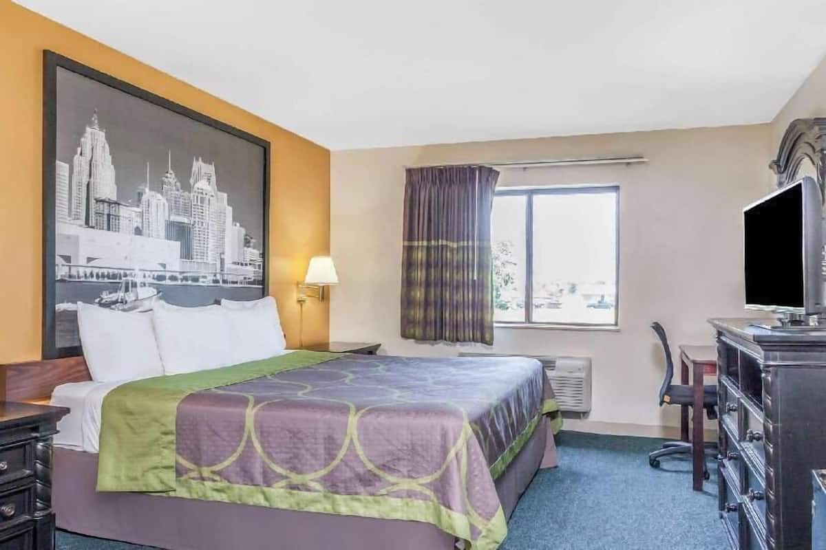 Ein Hotelzimmer mit Bett, Schreibtisch, Fernseher, einem Fenster mit Vorhängen und einem Stadtbild an der Wand.