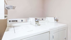 A row of Maytag washing machines with control panels, a white wall, and a door on the right. - Super 8 by Wyndham Imlay City (Imlay City)