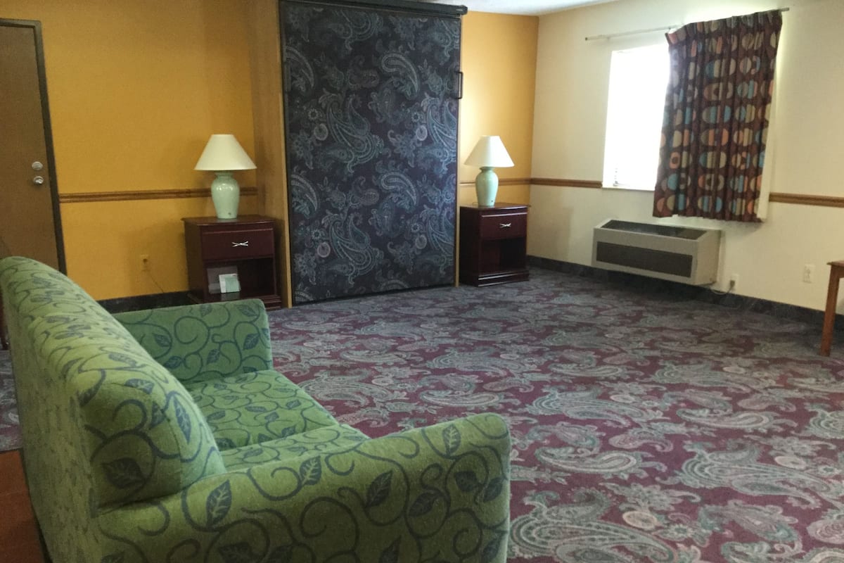 Ein Hotelzimmer mit Teppichmuster, zwei Nachttischen mit Lampen, einem großen Fenster mit Vorhängen und einer Tür.