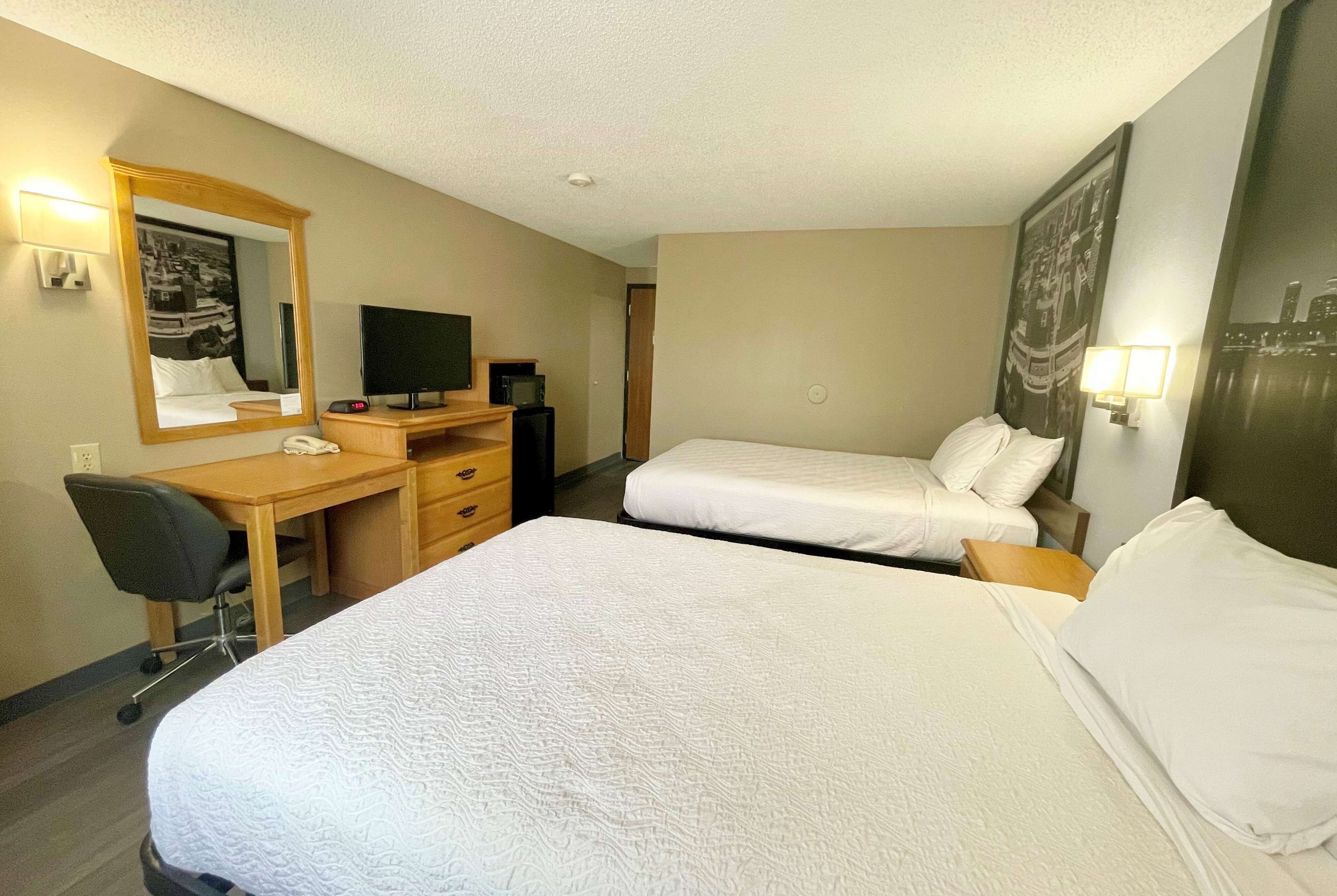 Quarto de hotel com duas camas, uma escrivaninha com computador, televisão e espelho.