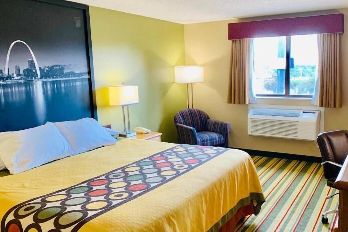 Een hotelkamer met een bed, een bureau, een stoel, een lamp, een airconditioner en een raam met gordijnen.