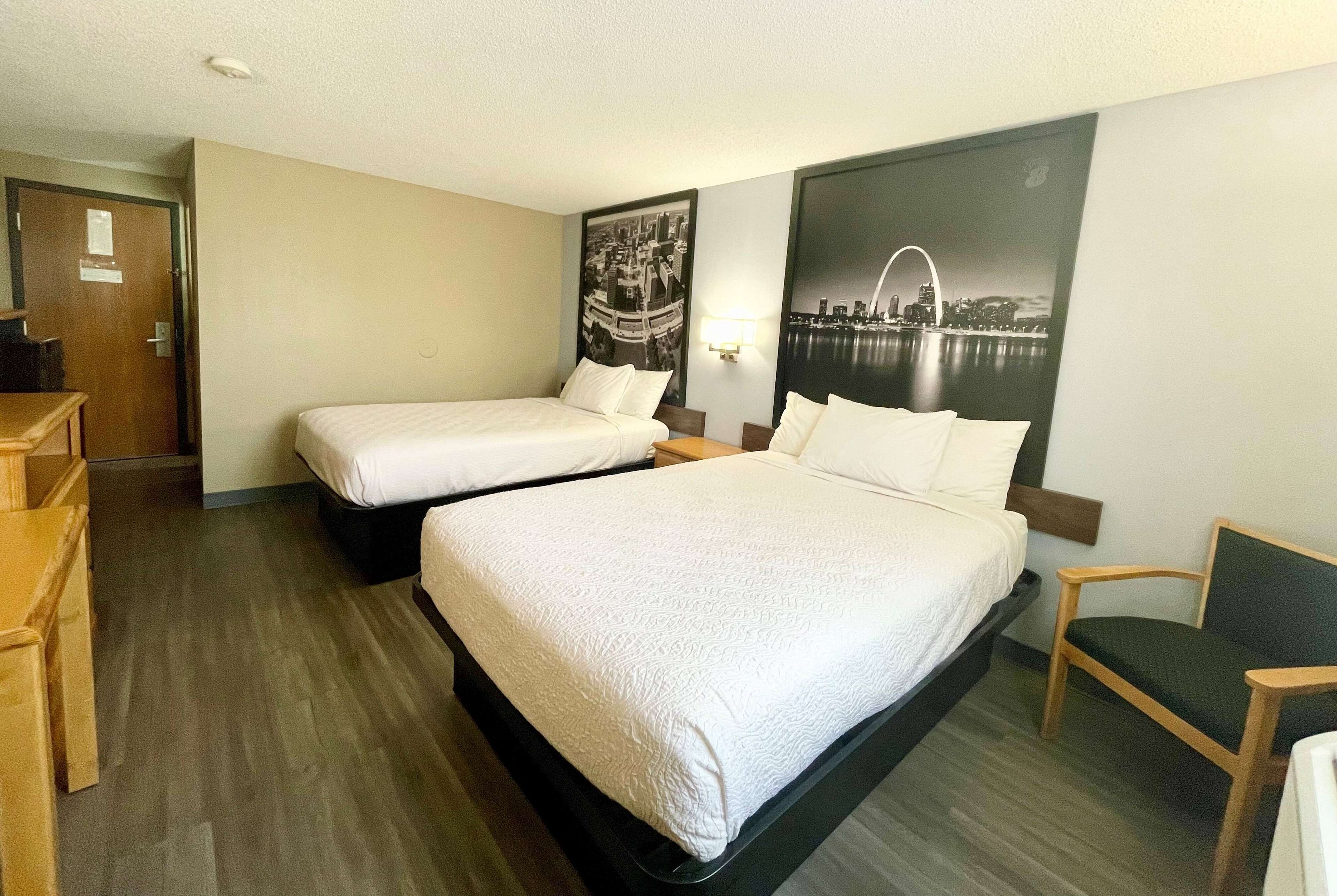 A hotel room with two beds, a wooden chair, a desk, and a cityscape painting on the wall.