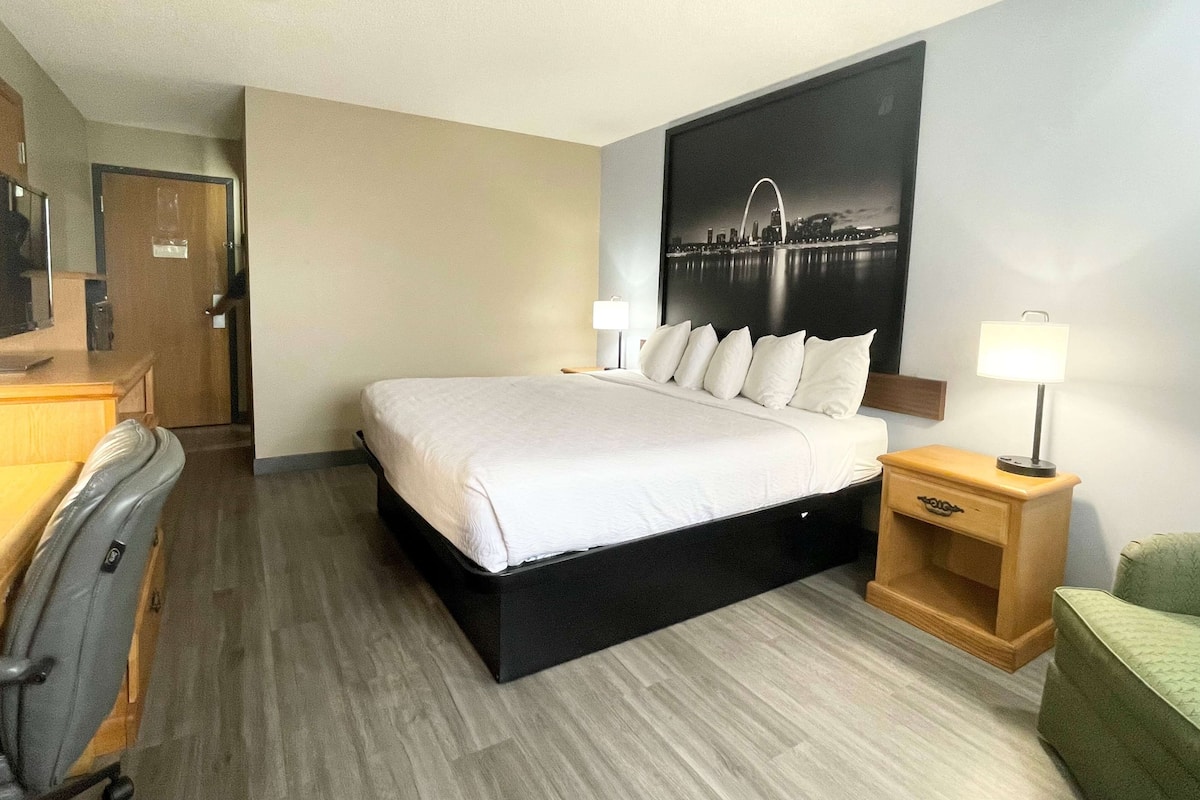 Een hotelkamer met een bed, nachtkastje, lamp, stoel, bureau en een stadsgezichtschilderij aan de muur.
