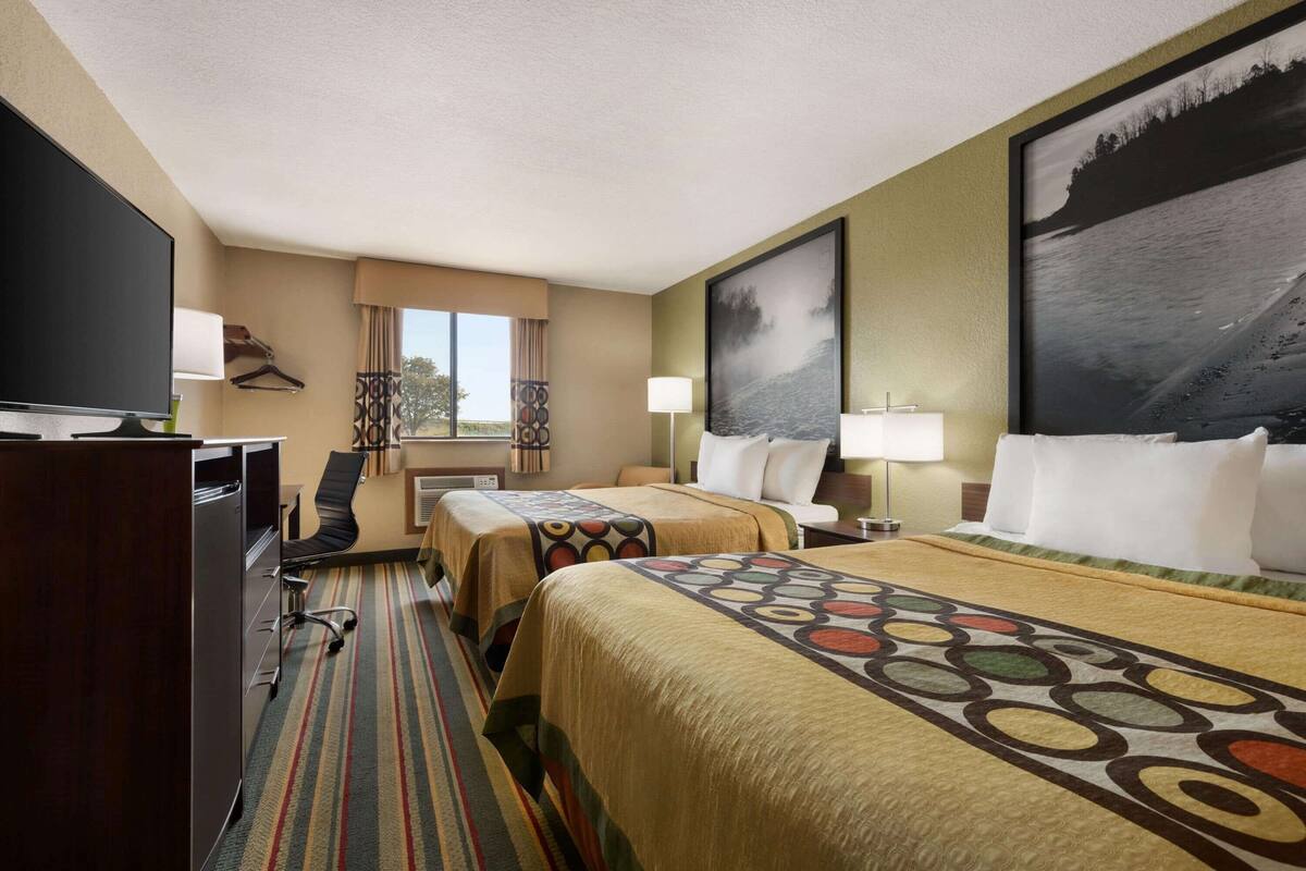 A hotel room with two beds, a desk with a TV, and a large window.