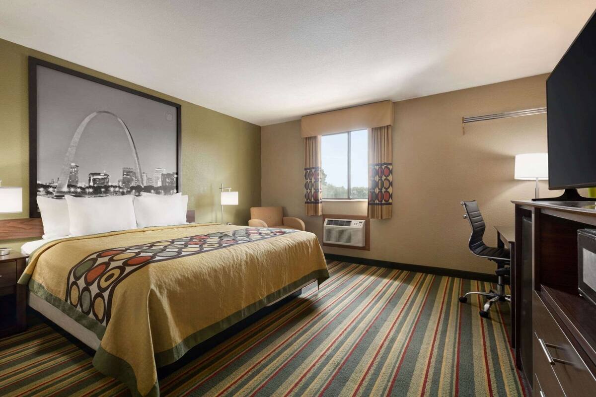 A hotel room with a bed, a desk, a chair, an air conditioning unit, and a television.