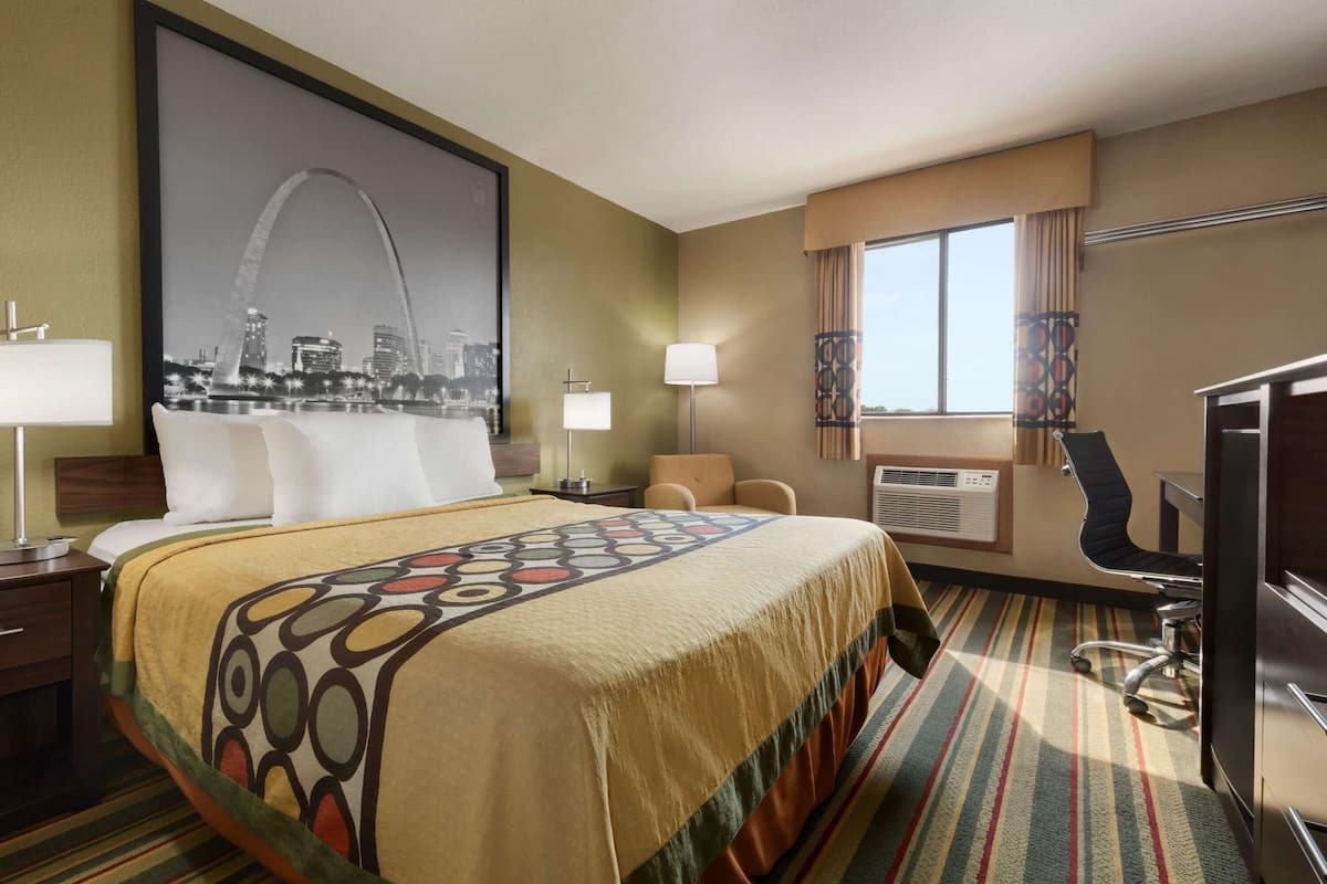 A hotel room with a large bed, a desk, a chair, a desk lamp, a window with curtains, and a wall-mounted air conditioning unit.