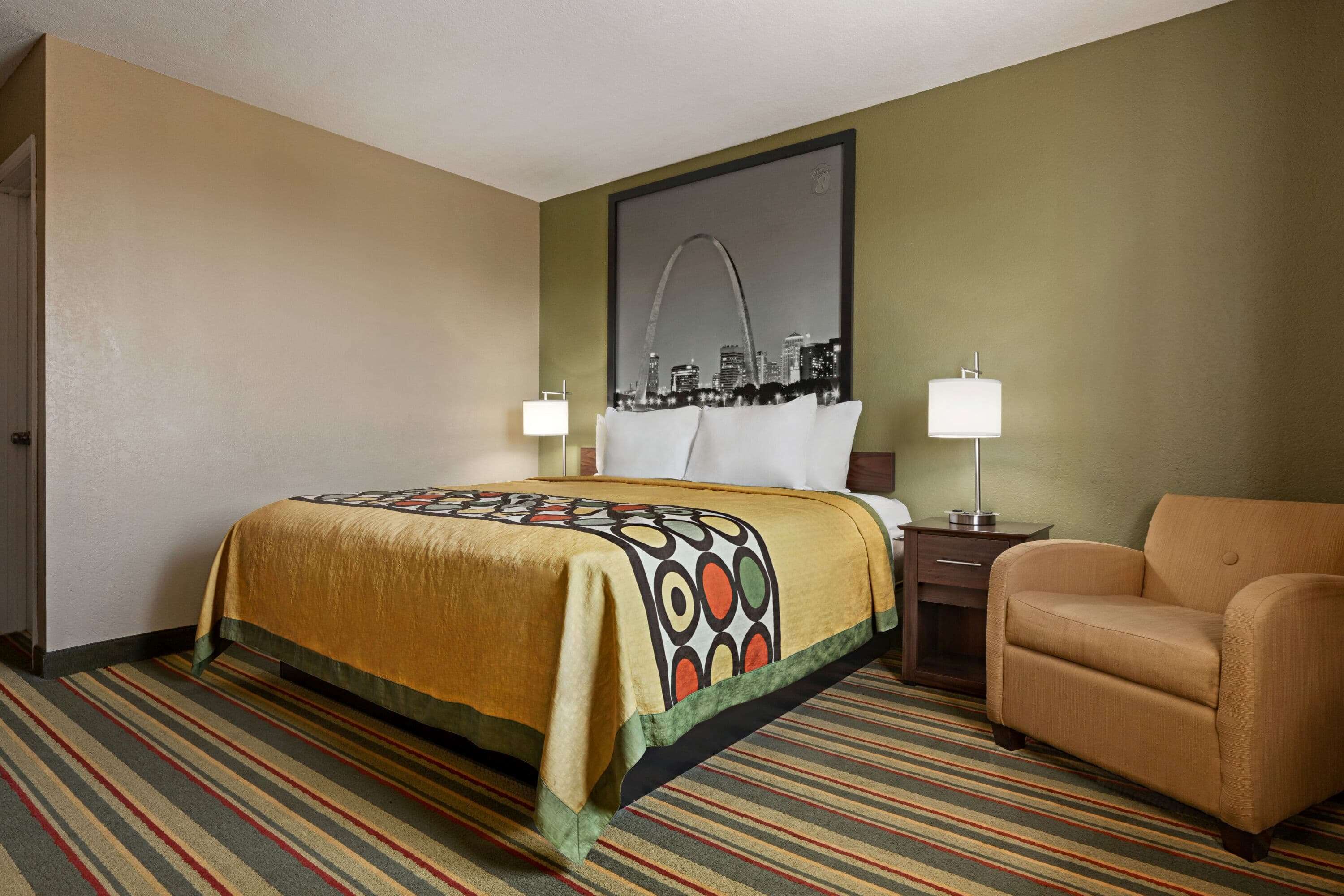 A hotel room with a bed, a chair, a nightstand, a lamp, and a cityscape painting on the wall.