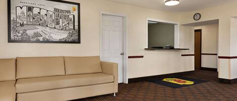 A hotel lobby with a beige sofa, a framed welcome sign, a reception desk, and a patterned carpet.
