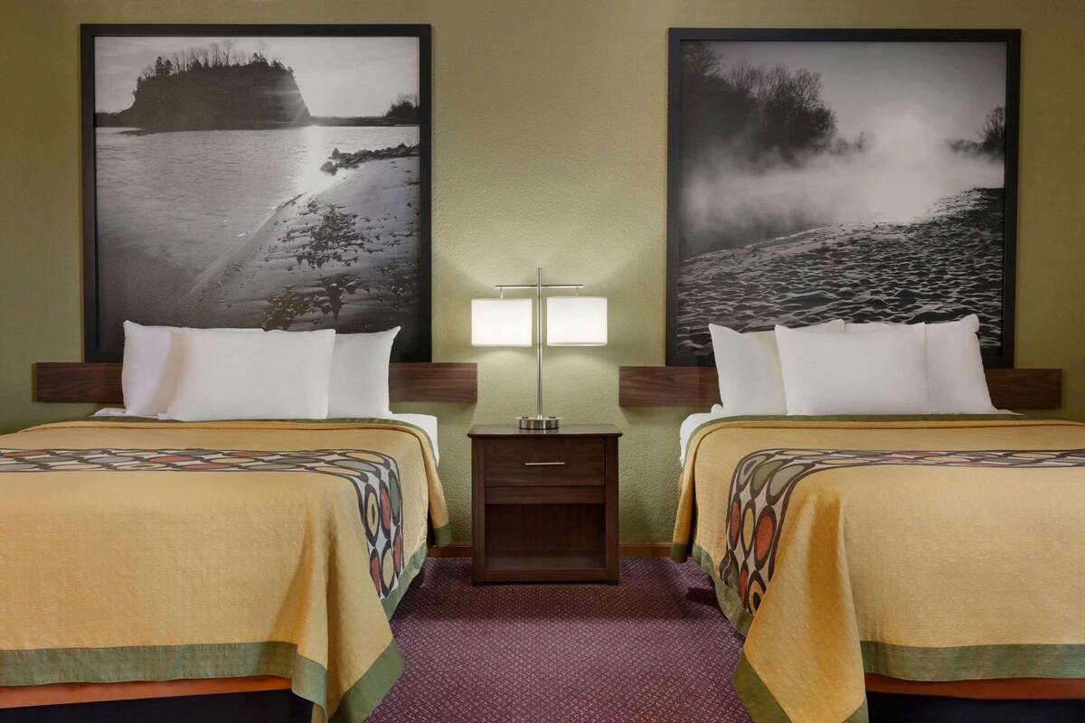 A hotel room with two beds, a nightstand with a lamp, and two large framed pictures of a landscape.